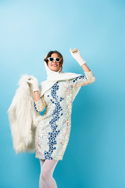 joyful woman in sunglasses and dress holding faux fur coat and gesturing on blue