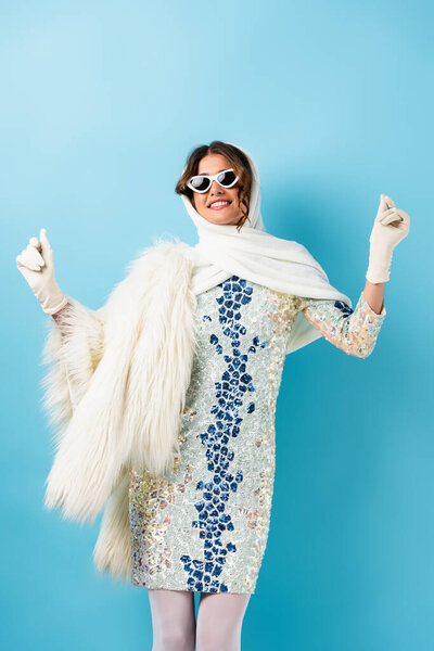 pleased woman in sunglasses and dress gesturing on blue