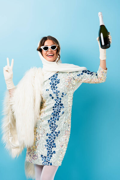 stylish woman in sunglasses smiling while holding bottle of champagne and showing peace sign on blue