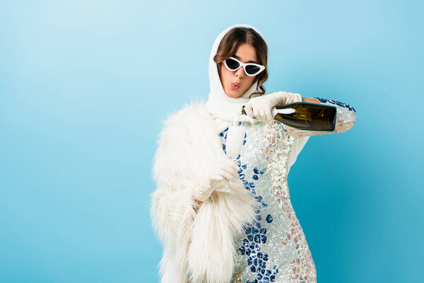 stylish woman in sunglasses pouring champagne in glass and whistling on blue