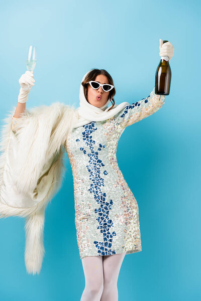 stylish woman in sunglasses and dress holding bottle of champagne and glass while whistling on blue