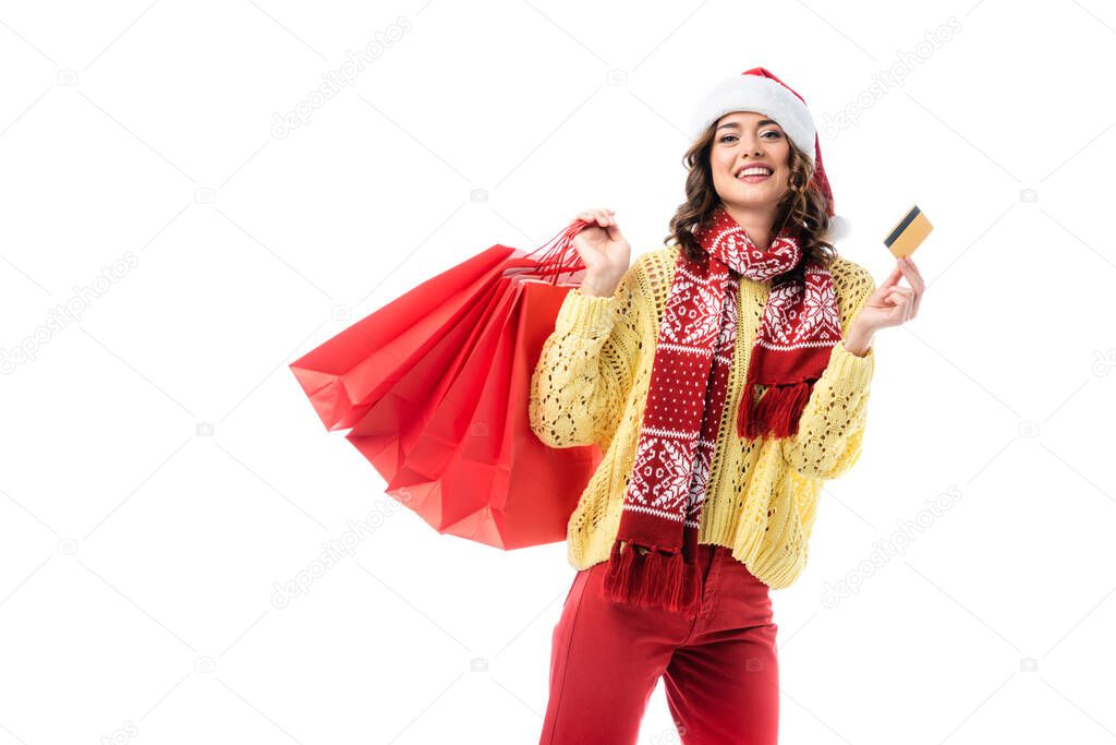 joyful woman in santa hat and scarf with ornament holding shopping bags and credit card isolated on white 
