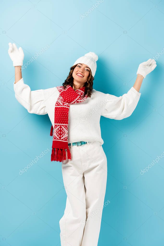 joyful woman in winter outfit, warm scarf, gloves and hat standing with outstretched hands on blue