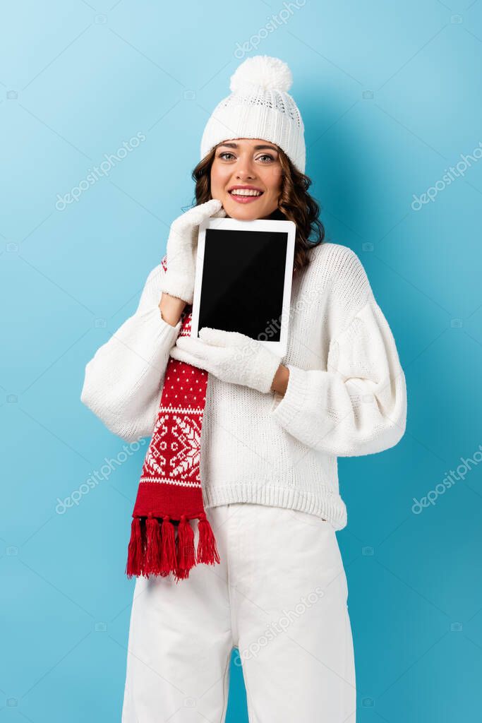 pleased woman in winter outfit holding digital tablet with blank screen on blue