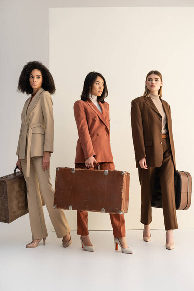 full length of young multicultural women in trendy suits holding suitcases on white