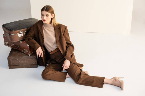 young woman in trendy suit leaning on luggage while posing on white