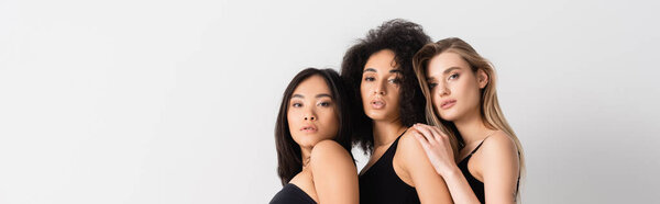 молодые межрасовые женщины в топах смотрят в камеру и позируют изолированно на белом, баннер