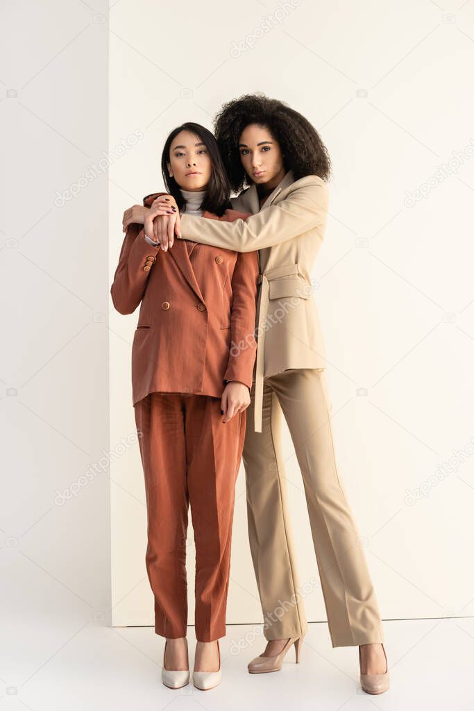 full length of african american and asian women in suits posing on white
