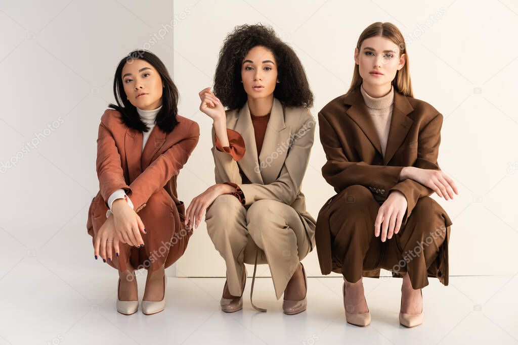 interracial young women in trendy suits sitting and posing on white