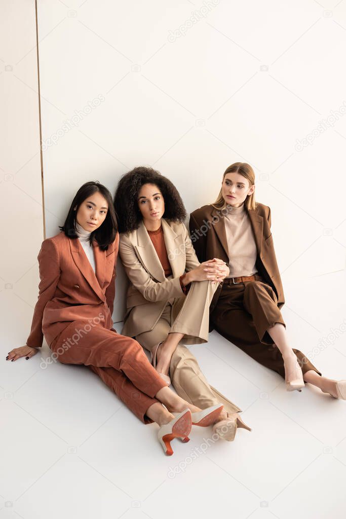 young multicultural women in trendy suits sitting and posing on white