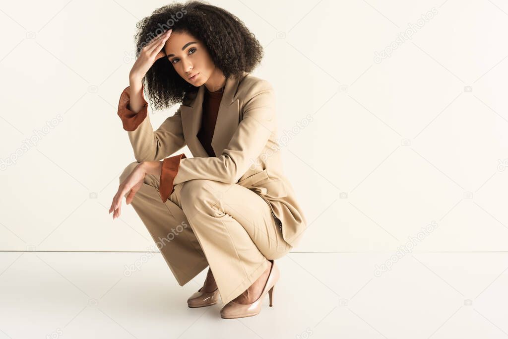 young and curly african american woman in stylish suit sitting on white