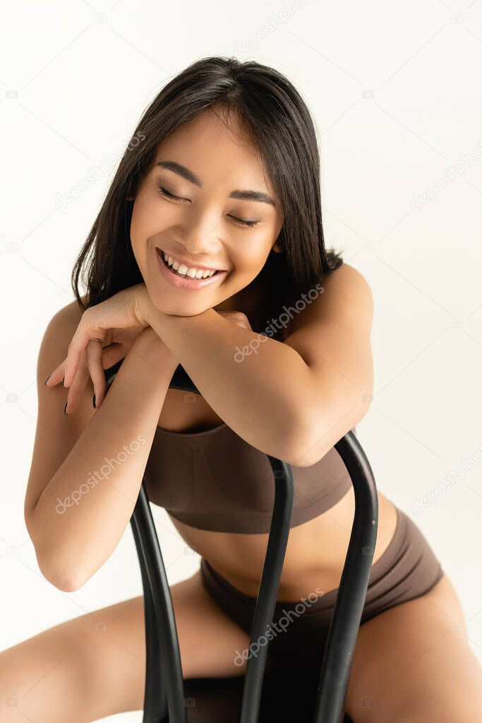 brunette asian woman in beige underwear smiling while posing on chair isolated on white