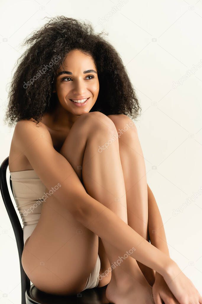 curly african american woman in underwear smiling while posing on chair isolated on white