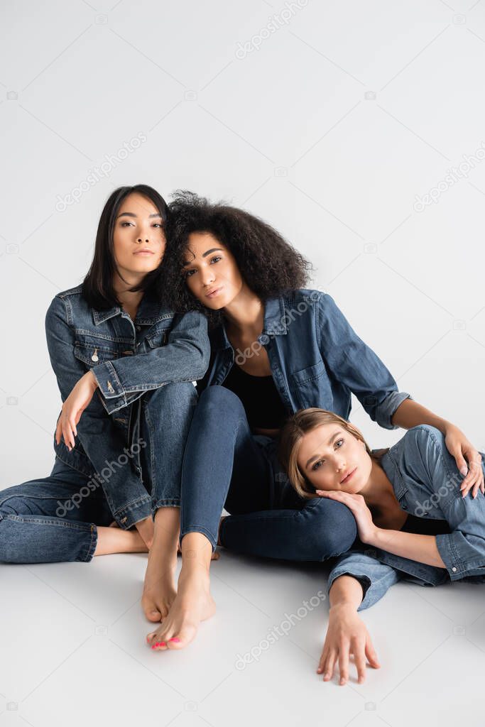 barefoot and interracial women in denim outfit posing on white