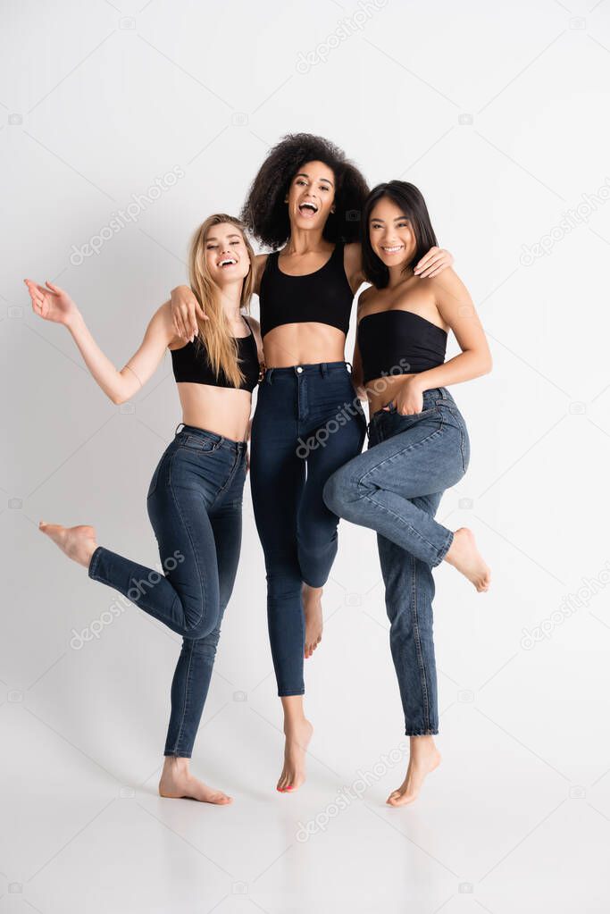 barefoot interracial women in denim jeans smiling while posing on white