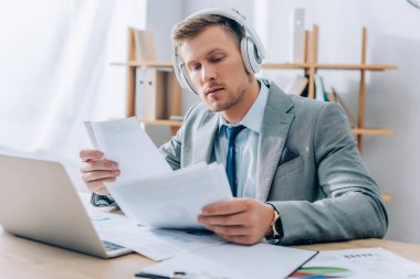 Kulaklıkla müzik dinleyen genç işadamı laptopun yanında ön planda bulanık kağıtlarla çalışırken 