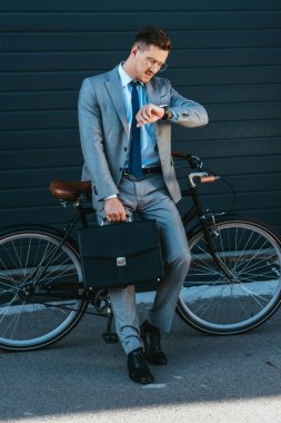 İş adamı dışarıda bisikletin yanında kol saatine bakarken evrak çantası tutuyor. 