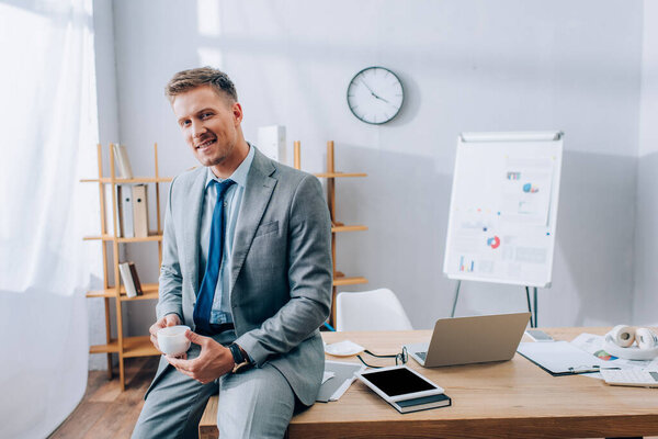 Cheerful businessman holding coffee cup near devices and papers in office 