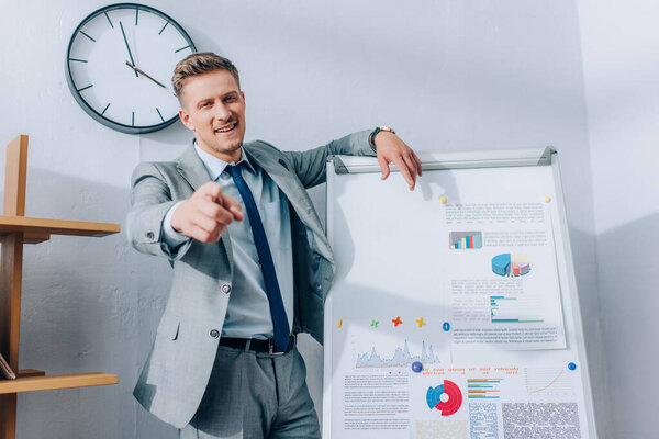 Cheerful businessman pointing at camera while standing near flipchart with graphs 