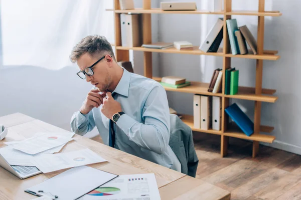 Üzletember Szemüveges Megérinti Nyakkendő Miközben Nézi Papírmunka Közelében Laptop Elmosódott — Stock Fotó