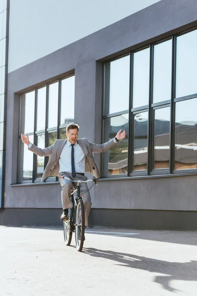 Cheerful businessman in formal wear riding bicycle without hands near building