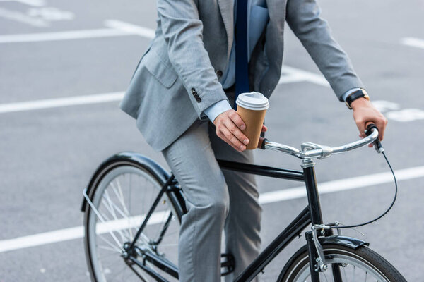 Cropped view of businessman holding takeaway coffee while riding bicycle outdoors 