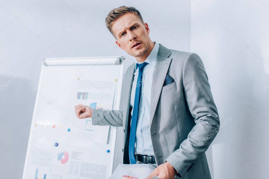 Focused businessman looking at camera while pointing at flipchart on blurred background in office 