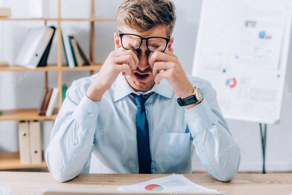 Businessman in eyeglasses rubbing tired eyes near papers on table