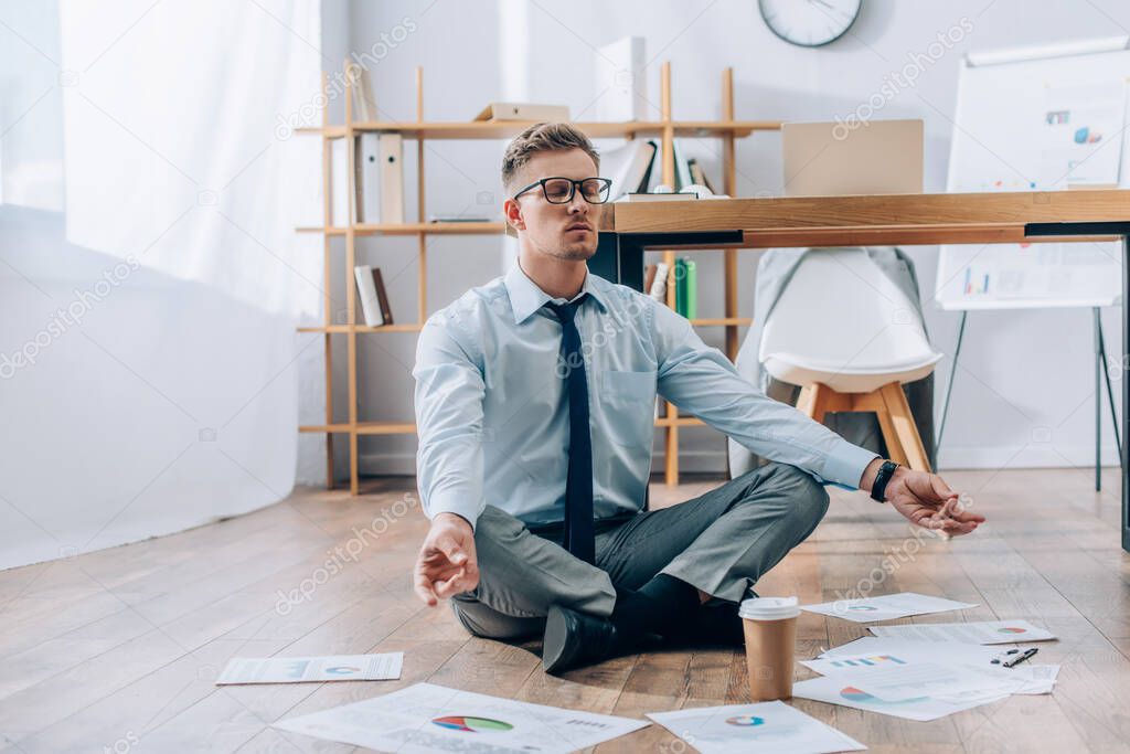 Young businessman meditating near papers and coffee to go on floor in office 