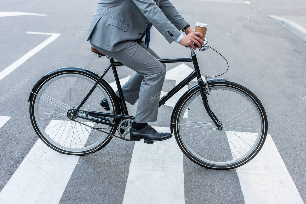 Cropped view of businessman holding takeaway coffee while cycling on crosswalk 