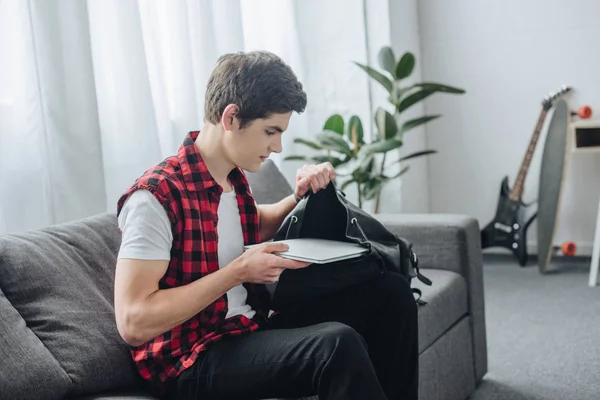 Adolescente masculino poner el ordenador portátil en el bolso mientras está sentado en el sofá - foto de stock