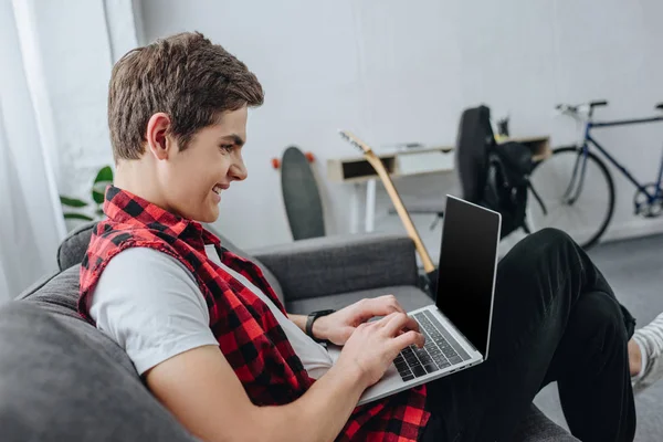 Sorrindo adolescente digitando no laptop enquanto sentado no sofá — Fotografia de Stock