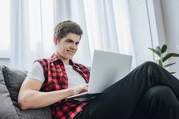 Lächelnder Teenie-Junge tippt auf Laptop, während er auf Sofa sitzt — Stockfoto