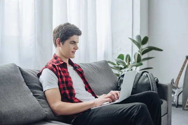 Beau adolescent tapant sur ordinateur portable tout en étant assis sur le canapé — Photo de stock