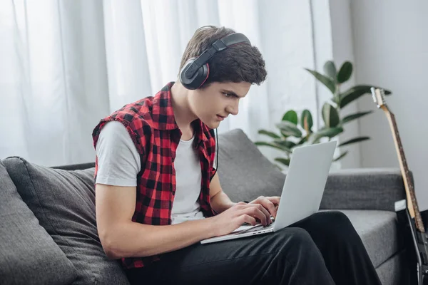 Concentrated teenager with headphones playing game on laptop while sitting on sofa — Stock Photo