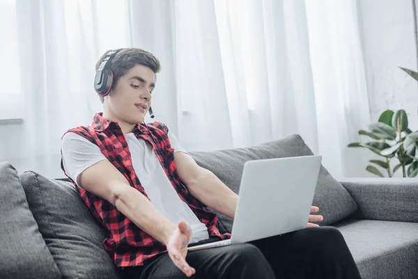 Хлопчик-підліток з навушниками грає в гру на ноутбуці, сидячи на дивані — стокове фото