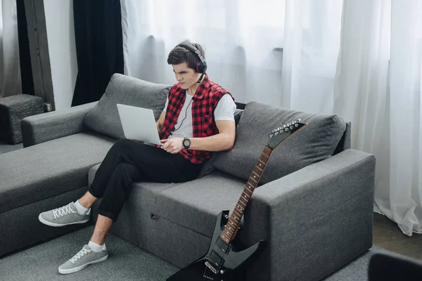 Teenager with headphones playing game on laptop while sitting on sofa with electric guitar near — Stock Photo
