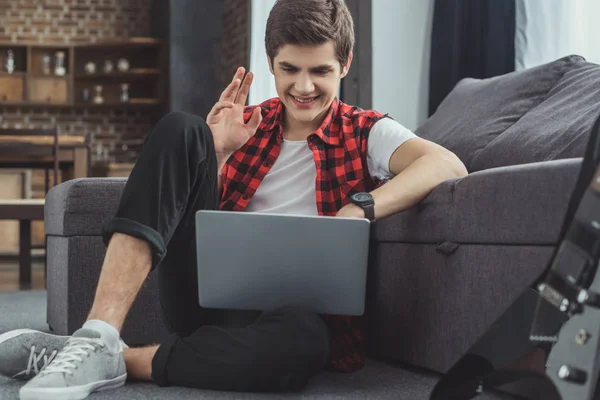 Adolescente sonriente haciendo videollamada en el portátil mientras está sentado en el piso cerca del sofá - foto de stock