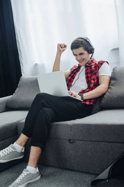 Веселий підліток з навушниками грає в гру на ноутбуці, сидячи на дивані — стокове фото