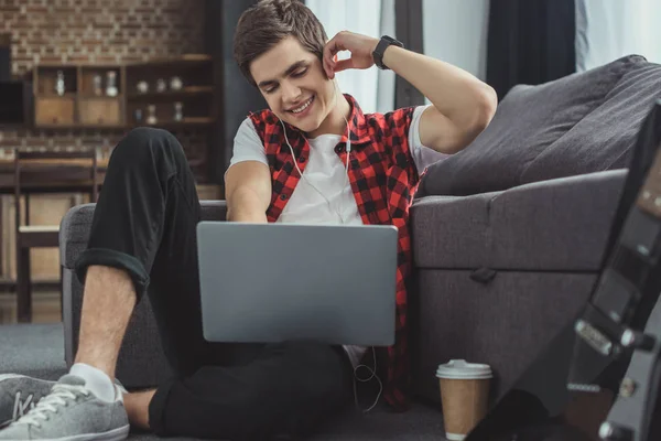 Handsome smiling teenager listening music with earphones and using laptop — Stock Photo