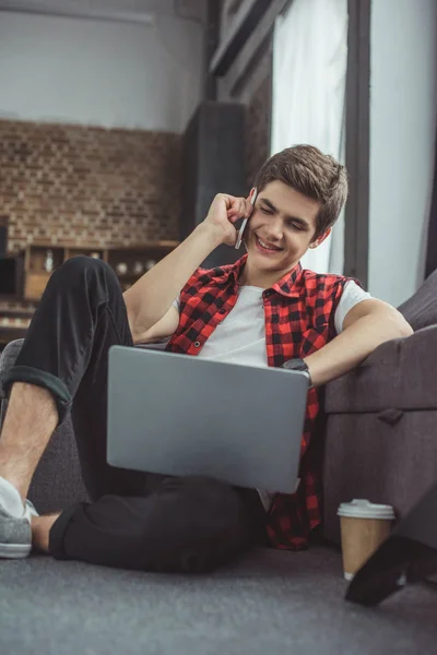 Adolescente alegre hablando en el teléfono inteligente y usando el ordenador portátil - foto de stock