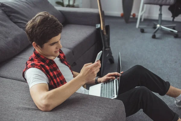 Handsome teenager using smartphone and laptop while sitting on floor — Stock Photo