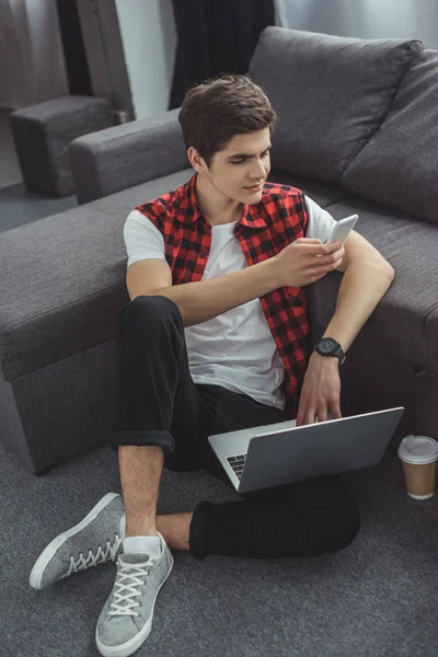 Улыбающийся подросток с помощью смартфона и ноутбука, сидя на полу с кофе — стоковое фото