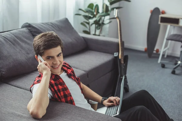Handsome smiling teenager talking on smartphone and using laptop — Stock Photo