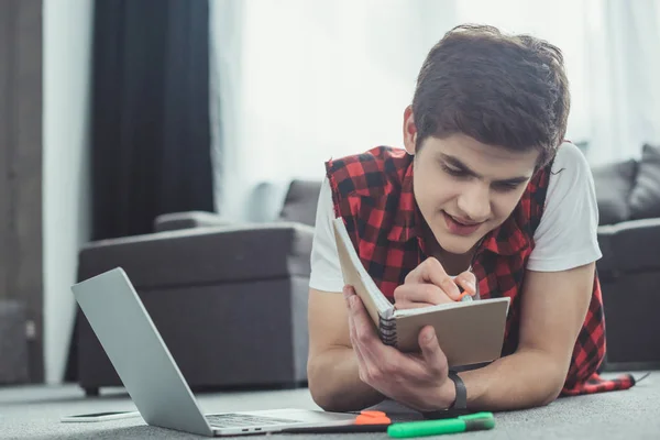 Handsome teenager studying with copybook and laptop while lying on floor — Stock Photo