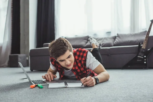 Хлопчик-підліток навчається з копіювальною книгою та ноутбуком, лежачи на підлозі — стокове фото