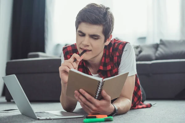 Pensativo adolescente estudiando con copybook y portátil mientras está acostado en el suelo - foto de stock