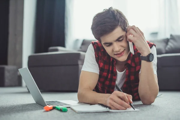 Guapo adolescente hablando en el teléfono inteligente mientras hace la tarea y tumbado en el suelo - foto de stock