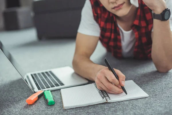 Vista recortada de adolescente hablando en el teléfono inteligente mientras hace la tarea y tumbado en el suelo — Stock Photo