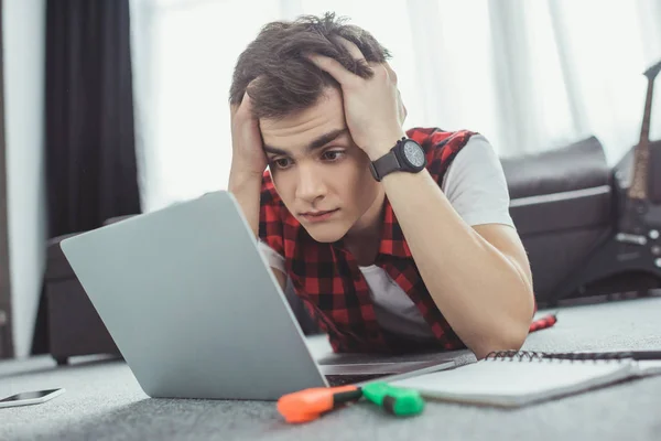 Stressed teen boy studying with laptop while lying on floor — Stock Photo